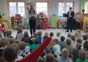 Dzieci ze wszystkich grup siedzą na dywanie, a przed nimi Pan prowadzący zajęcia opowiada bajkę o Elfince Klarze. Z tyłu stoi Pan z klarnetem. W tle okna, kąciki zabaw.