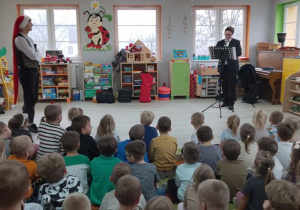 Dzieci siedzą na dywanie, a obok stoi Pan prowadzący zajęcia w czapce mikołajkowej. Z prawej strony stoi muzyk, który gra na klarnecie.