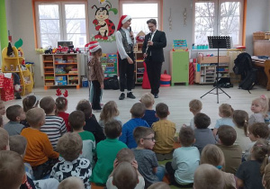 Dzieci siedzą na dywanie. Na scenie stoi troje dzieci w czapkach mikołajkowych, a obok nich Panowie omawiają budowę klarnetu.