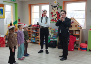 Troje dzieci stoi w czapkach mikołajkowych na scenie, a obok stoją zaproszeni goście. Pan gra na stroiku.