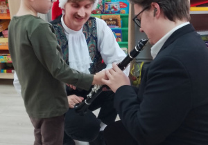 Mikołaj w czapce świątecznej dotyka klapki klarnetu. Obok przykucnęli zaproszeni goście.