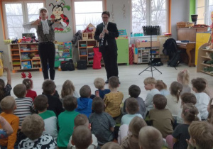 Dzieci siedzą na dywanie. Na środku sceny Pan gra na klarnecie piosenkę świąteczną. Obok niego stoi Pan prowadzący zajęcia.