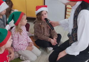 Grupka dzieci siedzi na dywanie, a obok przykucnął z mikrofonem Pan prowadzący zajęcia. Weronika śpiewa do mikrofonu kolędę.