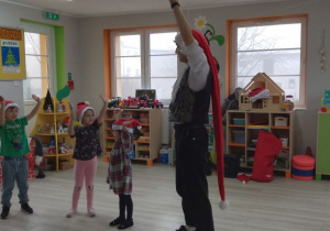 Troje dzieci bawi się na scenie razem z Panem prowadzącym zajęcia. Przedszkolaki podnoszą ręce do góry i grają na dzwonkach. W tle okna, kąciki zabaw.