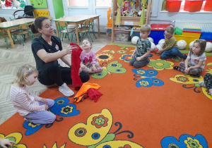 "Motylki" siedzą razem z Panią na dywanie, która pokazuje przedszkolakom znaczniki sportowe.