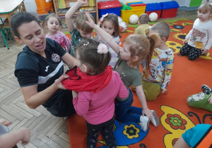 Pani zakłada dziewczynce znacznik sportowy w kolorze czerwonym.