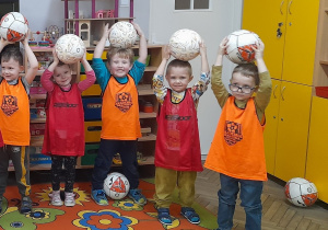 Pięciu chłopców ubranych w znaczniki sportowe stoi obok siebie z piłkami podniesionymi do góry.