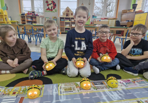 Oliwia, Wojtuś, Julek, Antoś i Tymek siedzą na dywanie z pszczółkami Bee Bot w dłoniach. Przed nimi leży mata do kodowania "droga".