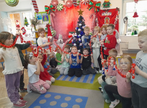 Dzieci trzymają wykonany przez siebie łańcuch w kolorze czerwonym. W tle choinka i dekoracja świąteczna.