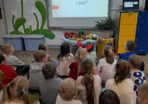 Dzieci siedzą przed tablicą multimedialną, na której wyświetlony jest pierwszy slajd prezentacji pt. "Choinka dawniej i dziś".