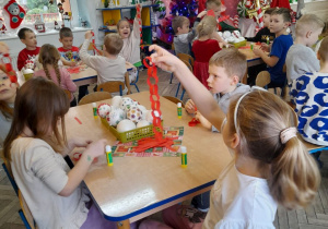 Dzieci siedzą przy stołach i wykonują łańcuch sklejając paski papieru w kolorze czerwonym. Na środku stołów leżą bombki w pojemnikach.