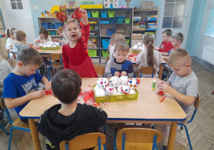 Dzieci przy stołach sklejają paski papieru w kolorze czerwonym. Tosia podniosła do góry wykonany przez siebie łańcuch.