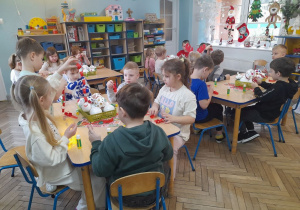 Dzieci siedzą przy stołach i wykonują łańcuch na choinkę. Na środku stołów leżą bombki w pojemnikach. W tle szafki z układankami, udekorowane świątecznie okno.