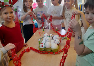 Grupka przedszkolaków stoi obok stołu. Dzieci połączyły wykonane przez siebie elementy łańcucha.