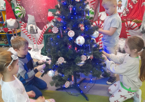 Czworo dzieci wiesza bombki na choince. W tle dekoracja świąteczna.