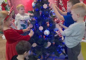 Pięcioro dzieci dekoruje choinkę bombkami. W tle dekoracja świateczna.