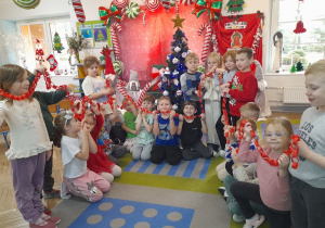 Dzieci trzymają wykonany przez siebie łańcuch w kolorze czerwonym. W tle choinka i dekoracja świąteczna.
