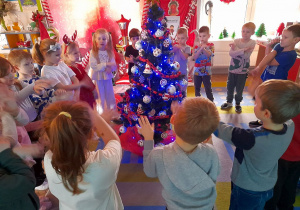 Dzieci stoją w kole na dywanie, a w środku ubrana, mieniąca się światełkami choinka. Przedszkolaki śpiewają piosenkę.