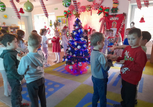 Dzieci bawią się na dywanie przy piosence. Na środku stoi ubrana choinka. W tle dekoracja świąteczna.