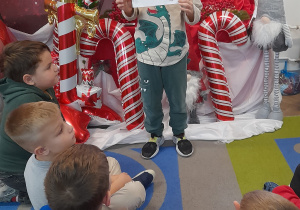 Fabian stoi przed tablicą, na której znajduje się kalendarz adwentowy. Chłopiec prezentuje kolejne zadanie z kalendarza, jakim jest ubieranie choinki.