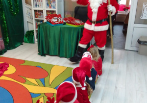 Pszczółki w mikołajowych czapkach siedzą na dywanie a ich wzrok skierowany jest na Mikołaja, który stanął w drzwiach sali, a w rękach trzyma złotą laskę i dzwonek.