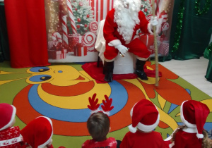 Mikołaj siedzi na honorowym miejscu na tle dekoracji z napisem Mikołajki a przed nim dzieci w mikołajkowych czapkach.