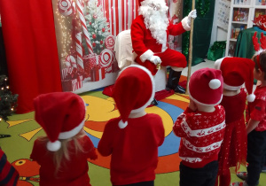 Mikołaj siedzi na honorowym miejscu a przed nim Pszczółki ubrane w czerwone stroje śpiewają piosenkę "Mikołaj". Z tyłu mikołajkowa dekoracja.