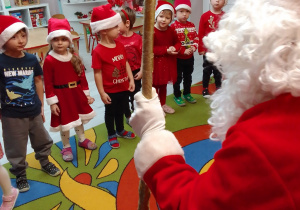 Dzieci w mikołajowych czapkach stoją w półkolu i prezentują piosenkę przygotowaną dla gościa. Na wprost dzieci siedzi Święty Mikołaj.