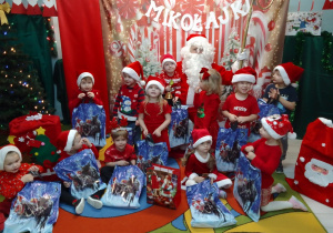 Grupa Pszczółek z prezentami pozują do zdjęcia z Mikołajem. W tle dekoracja mikołajkowa.