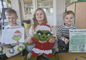 Na dywanie siedzą Wojtuś, Weronika i Olek. Wojtuś trzyma w dłoniach kopertę z wizerunkiem Grincha, Weronika filcową podobiznę skrzata, a Oluś list, który złośliwy skrzat wysłał do dzieci.