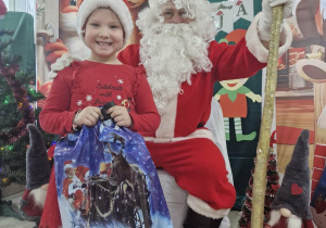 Majka stoi obok Świętego Mikołaja. Dziewczynka trzyma w dłoniach prezent.