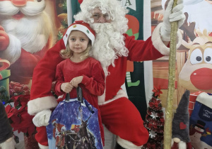 Liliana pozuje do zdjęcia ze Świętym Mikołajem na tle mikołajkowej dekoracji.