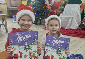 Maja i Ola siedzą na dywanie. Dziewczynki pokazują do zdjęcia kalendarze adwentowe otrzymane od Mikołaja.