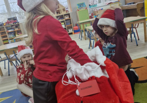 Alicja stoi przed dużym czerwonym workiem pozostawionym przez Świętego Mikołaja i wyjmuje z niego prezenty.