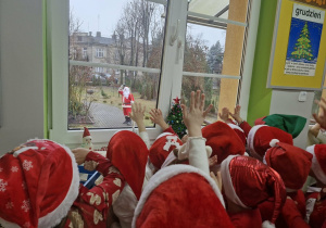 Biedronki stoją przy oknie, przez które widać idącego Mikołaja.