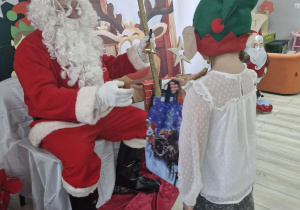 Święty Mikołaj siedzi na krześle ustawionym przed świąteczną dekoracją. Przed Mikołajem stoi Weronika ubrana w zielono – czerwoną czapkę elfa.