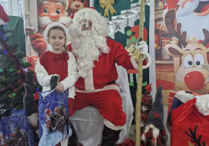 Ola stoi obok Świętego Mikołaja. Dziewczynka trzyma w dłoniach prezent.