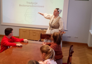 Grupka dzieci siedzi przy stole, przed nimi Pani ubrana w strój z XVIII wieku opowiada o tradycjach bożonarodzeniowych prezentując slajdy.