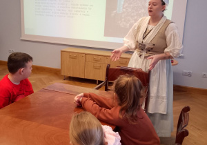 Pani ubrana w strój z XVIII wieku stoi przed dziećmi i rozmawia na temat Świąt Bożego Narodzenia.