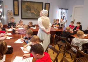 Przedszkolaki siedzą przy wspólnym stole i kolorują obrazki świąteczne. Obok stoi pani w stroju z XVIII wieku.