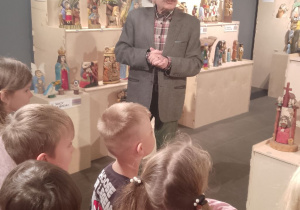 Pan rzeźbiarz stoi przed dziećmi na tle wystawy rzeźb.