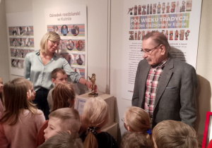 Pani etnograf pokazuje dzieciom nagrodę - statuetkę Lwa Kutnowskiego, obok stoi rzeźbiarz na tle plakatów - Ośrodek rzeźbiarski w Kutnie.
