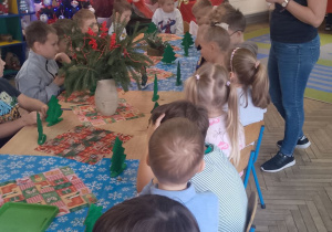 Dzieci siedzą przy wspólnym stole, obok stoi pani z Centrum Kultury Gminy Kutno i omawia kolejny etap prac - dekorowanie ozdobami.