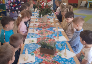 Dzieci siedzą przy wspólnym stole, obok stoi pani z Centrum Kultury Gminy Kutno. Na stole przed każdym dzieckiem stoi choinka ze sklejki oraz leżą pędzelki i gąbeczki.