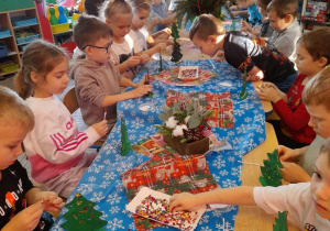 Dzieci siedzą przy wspólnym stole i ozdabiają choinki cekinami.