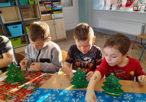 Trzech chłopców siedzi obok siebie przy stole i dekoruje choinki cekinami.