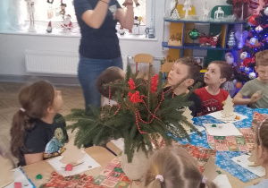 Pani pokazuje dzieciom wykonaną choinkę i omawia etapy jej przygotowania. Przedszkolaki siedzą przy stole.