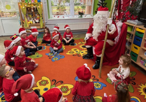 Dzieci siedzą w kole na dywanie, a Mikołaj siedzi na czerwonym krześle.