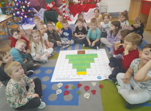 Dzieci siedzą na dywanie przed matą do kodowania, na której znajduje się ułożona choinka z zielonych kartoników. Obok maty leży kilka ozdób choinkowych.