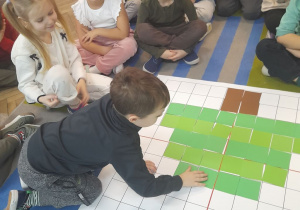 Bartek układa na macie do kodowania choinkę z zielonych kartoników według wzoru. Obok grupka dzieci obserwuje działanie kolegi.